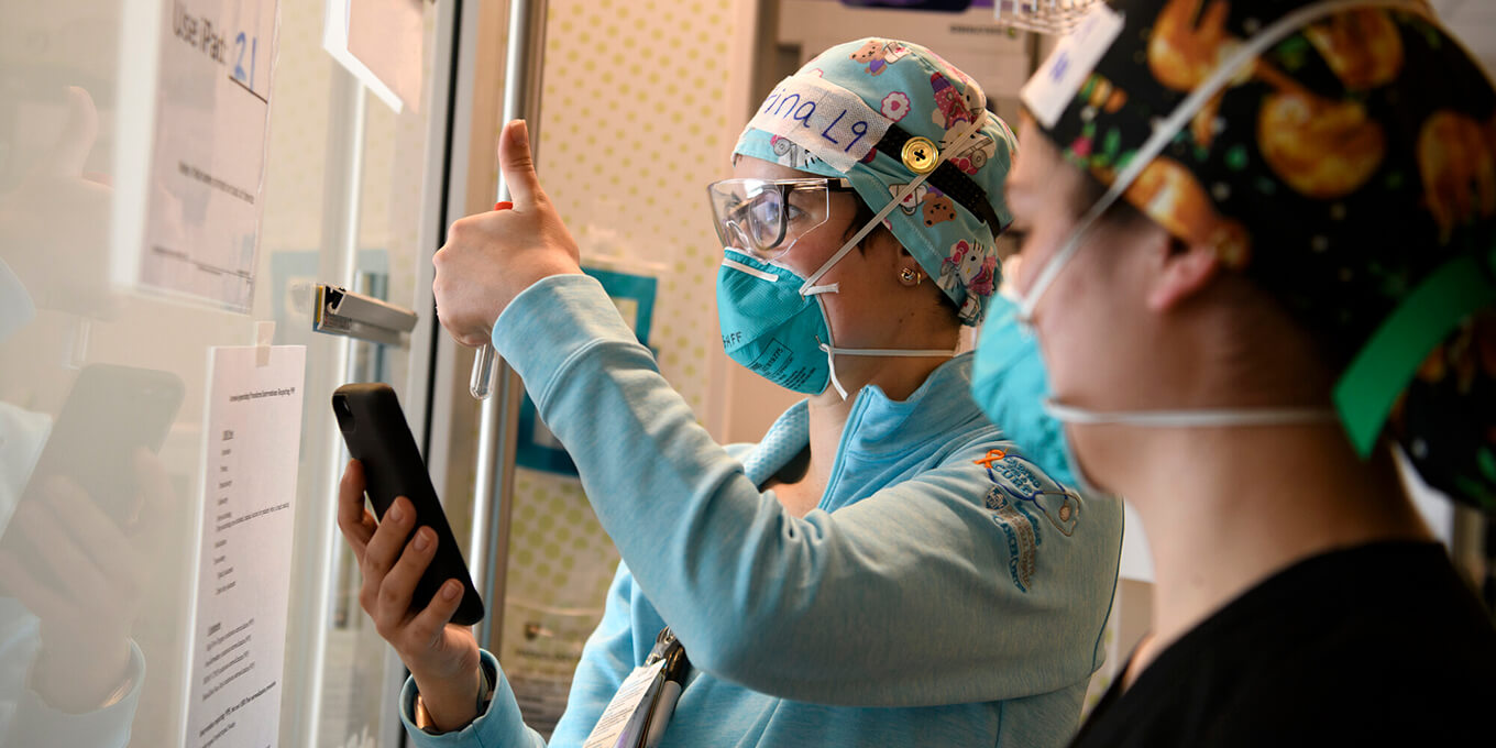 Two care providers in scrubs, caps and gloves look through a glass partition. One gives a thumbs up to someone on the other side.