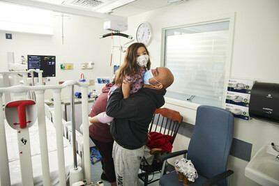 Inside of a hospital room, a man lifts up a child with long hair. They both have masks on.